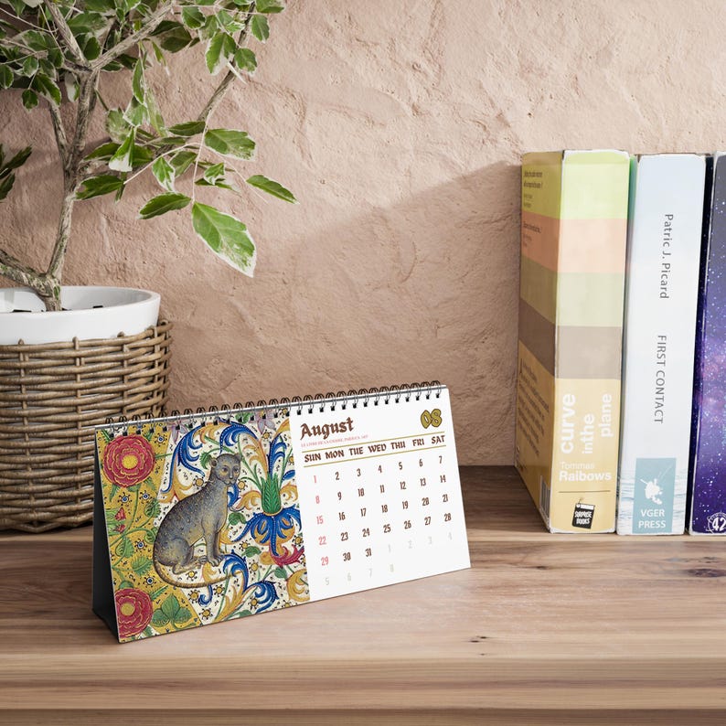 May include: A desk calendar for the month of August featuring a spiral binding at the top. The left side displays a colorful illustration of a cat surrounded by floral patterns, while the right side shows the calendar grid with the text August.