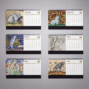 May include: A spiral-bound desk calendar displaying six months from January to June. Each page features a medieval-style illustration of a cat alongside a grid of dates. The months are labeled with text and numbers, with a black base at the bottom.