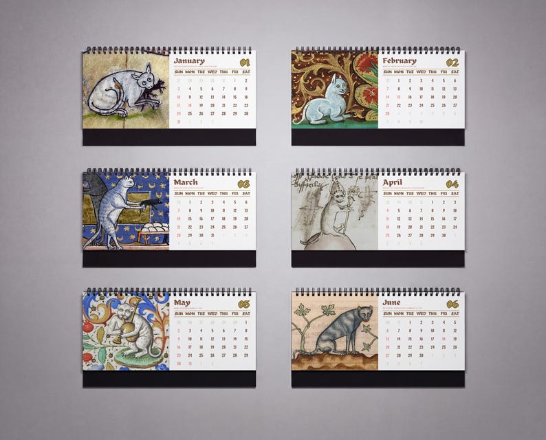 May include: A spiral-bound desk calendar displaying six months from January to June. Each page features a medieval-style illustration of a cat alongside a grid of dates. The months are labeled with text and numbers, with a black base at the bottom.
