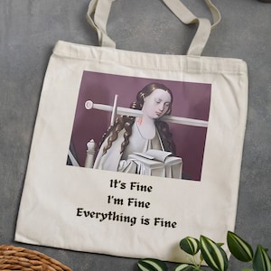 May include: Cream-colored tote bag featuring a medieval-style image of a woman with a sword and book. The text "It's Fine, I'm Fine, Everything is Fine" is printed below the image in a black, gothic font. The bag has two handles.