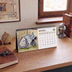 May include: A desk calendar featuring a spiral binding and a white page for January. The left side displays a medieval-style illustration of a grey cat-like creature, while the right side lists the days of the month in a dark brown font.
