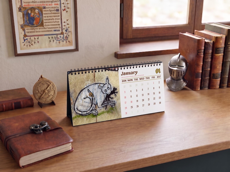 May include: A desk calendar featuring a spiral binding and a white page for January. The left side displays a medieval-style illustration of a grey cat-like creature, while the right side lists the days of the month in a dark brown font.