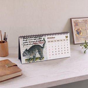 May include: A spiral-bound desk calendar featuring a medieval-style illustration of a grey cat on the left page and the month of October with a grid of dates on the right page. The calendar is displayed in a landscape orientation with black wire binding.