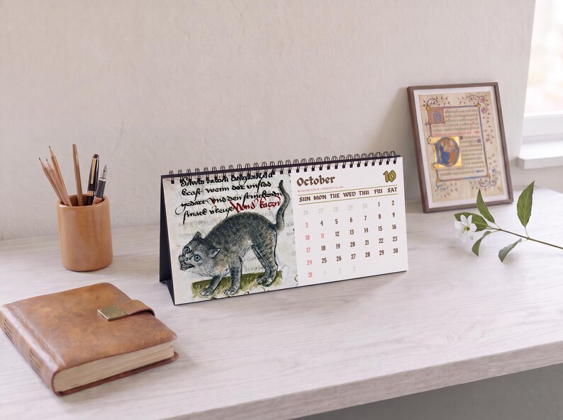 May include: A spiral-bound desk calendar featuring a medieval-style illustration of a grey cat on the left page and the month of October with a grid of dates on the right page. The calendar is displayed in a landscape orientation with black wire binding.