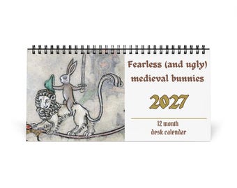 May include: A 2027 12-month desk calendar with a black spiral binding. The cover features a medieval-style illustration of a rabbit holding a sword and shield while riding a lion, with the text Fearless (and ugly) medieval bunnies 2027.