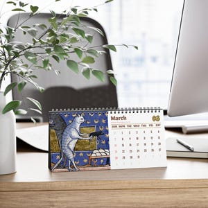 May include: A spiral-bound desk calendar for the month of March, featuring a medieval-style illustration of a cat holding a small creature against a blue background with gold stars and moons, alongside a grid of dates printed in black and red ink.
