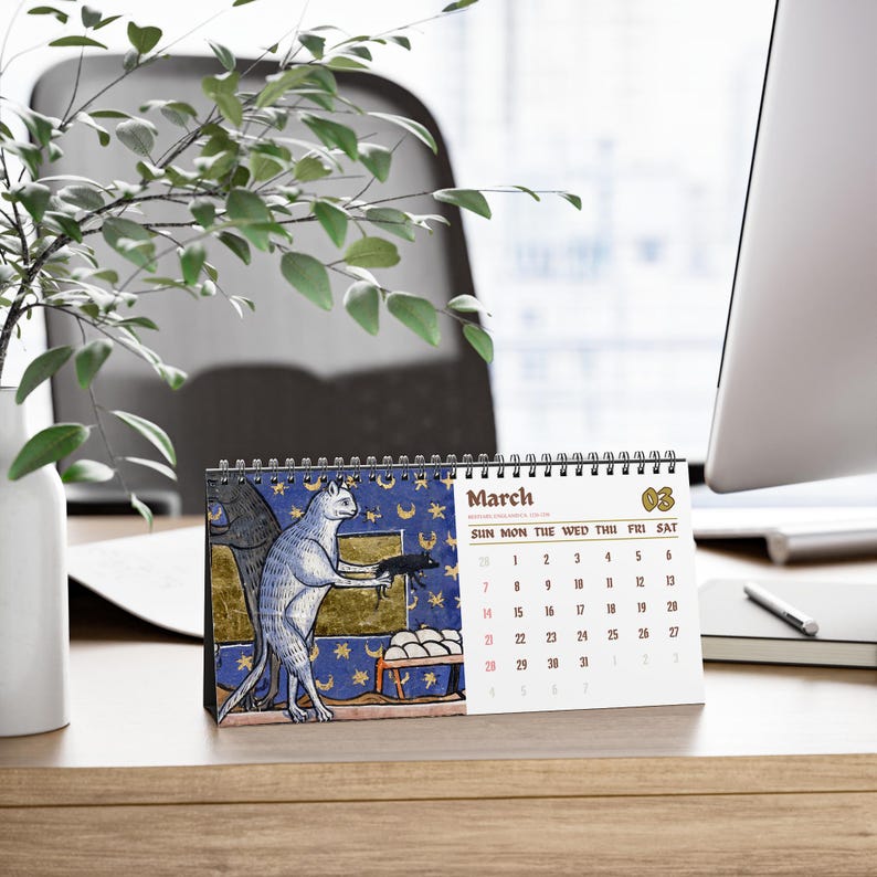 May include: A spiral-bound desk calendar for the month of March, featuring a medieval-style illustration of a cat holding a small creature against a blue background with gold stars and moons, alongside a grid of dates printed in black and red ink.