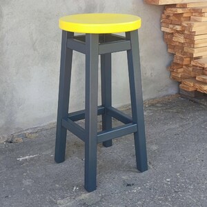 Op de afbeelding: Een hoge, grijze houten kruk met een ronde, felgele zitting. De kruk heeft vier poten en een voetensteun. De achtergrond is een grijze betonnen muur en een stapel houten planken.