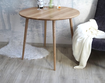 Puede incluir: Una mesa redonda de madera clara con tres patas. La mesa tiene una superficie lisa. Una alfombra blanca y esponjosa está en el suelo. Una manta blanca está sobre una silla gris oscura.