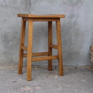 Könnte beinhalten: Ein Barhocker aus Holz mit quadratischer Sitzfläche und vier Beinen. Der Hocker ist aus hellbraunem Holz gefertigt und hat ein schlichtes, rustikales Design. Der Hocker ist etwa 61 cm hoch.