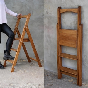 May include: A wooden step stool with two steps and a top shelf. The stool is folded and standing against a wall. The stool is made of natural wood and has a light brown finish.