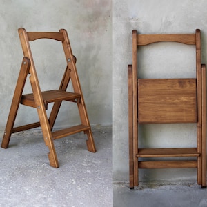 May include: A wooden folding step stool in a warm brown finish. The stool has a seat and two steps, with a curved top. The stool is shown both open and folded, showcasing its space-saving design. The wood grain is visible.