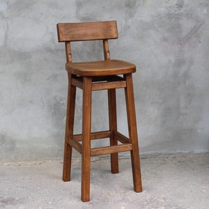 May include: A solid wood bar stool with a brown finish. The stool has a rectangular seat and a back rest. The stool has a simple design with four legs and a crossbar for support.