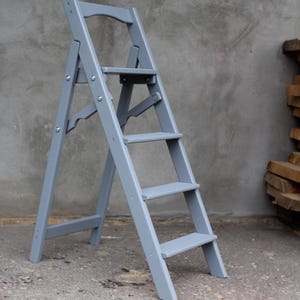 K&ouml;nnte beinhalten: Eine graue Holztreppe mit f&uuml;nf Stufen und einer kleinen Plattform oben. Die Leiter ist zusammengeklappt und steht auf einer Betonfl&auml;che.