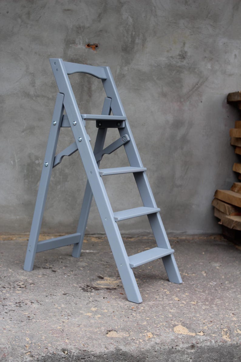 K&ouml;nnte beinhalten: Eine graue Holztreppe mit f&uuml;nf Stufen und einer kleinen Plattform oben. Die Leiter ist zusammengeklappt und steht auf einer Betonfl&auml;che.
