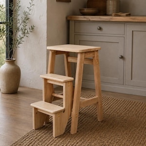 May include: A light-colored wooden step stool featuring three tiers. The stool has a rectangular top platform, a middle step, and a lower step, all supported by four sturdy legs. The natural wood grain is visible throughout the entire structure.