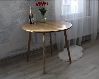 Puede incluir: Una mesa redonda de madera con un acabado marrón claro y tres patas delgadas. La mesa está hecha de roble macizo y tiene una veta de madera natural.