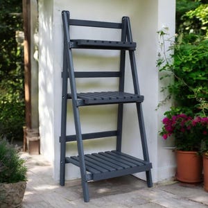 Op de afbeelding: Een grijze houten plantenstandaard met drie planken. De standaard leunt tegen een witte muur en heeft een rustieke uitstraling.