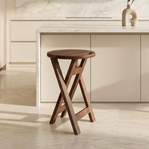May include: A dark brown wooden stool with a circular seat and a folding X-frame base. The seat features a small integrated handle cutout on one side, and the legs are connected by a horizontal support bar, creating a sturdy, collapsible structure.