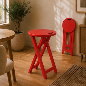 May include: A bright red folding stool with a circular seat and a cutout handle. The stool features a cross-leg base design. A second identical red stool is shown in a folded, upright position in the background, displaying its circular top.