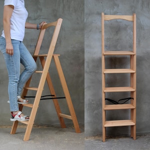 Könnte beinhalten: Eine hellbraune Holztrittleiter mit einem Klappdesign. Die Leiter hat fünf Regale und wird von einer Person benutzt. Die Leiter kann als Trittschemel oder kleines Bücherregal verwendet werden. Die Leiter ist etwa 150 cm hoch.