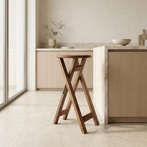 Puede incluir: Un taburete alto plegable de madera con asiento circular y base de patas cruzadas. El taburete está construido con madera de tono medio con un acabado liso, con cuatro patas que se cruzan para proporcionar soporte y estabilidad.