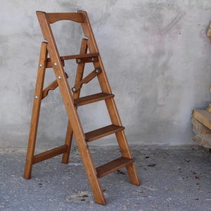 K&ouml;nnte beinhalten: Eine Holztreppe mit vier Stufen. Die Leiter ist aus Naturholz gefertigt und hat eine braune Oberfl&auml;che. Die Leiter ist zusammengeklappt und einsatzbereit.