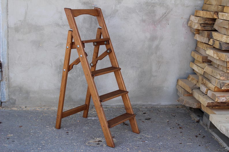 K&ouml;nnte beinhalten: Eine Holztreppe mit vier Stufen. Die Leiter ist aus Naturholz gefertigt und hat eine braune Oberfl&auml;che. Die Leiter ist zusammengeklappt und einsatzbereit.