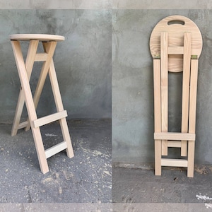 Könnte beinhalten: Ein klappbarer Holzhocker mit runder Sitzfläche und schlichtem Design. Der Hocker ist aus Naturholz gefertigt und hat eine hellbraune Farbe. Der Hocker ist zusammengeklappt und steht selbstständig.