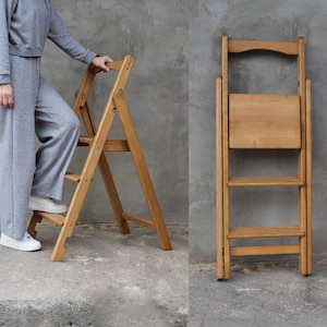 May include: A wooden folding step stool in natural wood tones. The stool is shown both open and folded, with a person using the steps. The stool has three steps and a small backrest when open. The wood grain is visible.