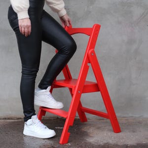 May include: A bright red, two-step folding step stool. The stool is made of wood and has a simple design. The step stool is shown in front of a gray wall. The stool is approximately 20 inches tall.