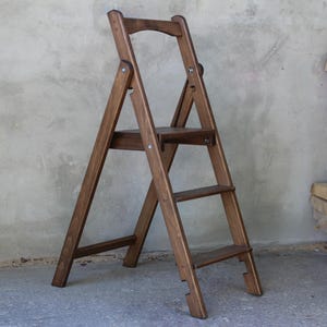 Könnte beinhalten: Ein klappbarer Trittschemel aus Holz in Dunkelbraun. Er hat drei Stufen und einen gebogenen Griff. Das schlichte Design ist ideal zum Erreichen höherer Stellen. Die Höhe beträgt etwa 90 cm.