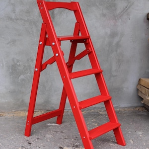 K&ouml;nnte beinhalten: Eine rote Holztreppe mit f&uuml;nf Stufen. Die Treppe ist zusammengeklappt und steht auf einer Betonfl&auml;che.