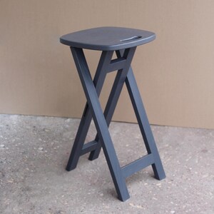 May include: A dark grey folding stool with a wooden top and a handle. The stool is made of wood and has a simple, modern design.