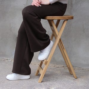 May include: A wooden folding stool with a round seat. The stool has an X-frame design and is made of light-coloured wood. A person is sitting on the stool, wearing brown trousers and white trainers.