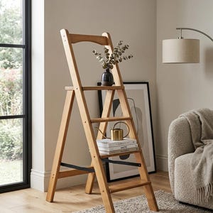 Könnte beinhalten: Eine Holz-Trittleiter mit einem Regal, auf dem Bücher, eine Gießkanne und eine Vase mit Zweigen stehen. Die Leiter ist hellbraun und hat eine schwarze Metallstütze. Der Raum hat eine neutrale Farbpalette.