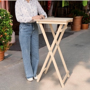 Puede incluir: Una mesa plegable de madera clara con una tapa rectangular y un diseño simple y robusto. La mesa está sobre una superficie de hormigón.