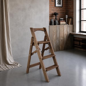 May include: A folding wooden step stool with three steps and a curved top handle. The stool features a medium brown wood grain finish, metal hinges, and silver-coloured hardware connecting the frame and steps in a sturdy, functional design.