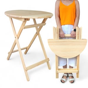 Op de afbeelding: Een lichtgekleurde, houten klaptafel met een rond blad en een bijpassende klapstoel. De tafel heeft een diameter van ongeveer 61 cm en is 71 cm hoog wanneer hij open is. De stoel is opgevouwen en wordt door een persoon vastgehouden.