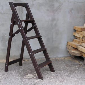 K&ouml;nnte beinhalten: Eine dunkelbraune Holz-Treppe mit vier Stufen. Die Leiter ist zusammengeklappt und steht auf einem Betonboden.