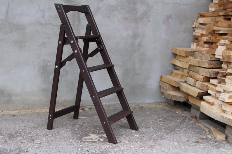 K&ouml;nnte beinhalten: Eine dunkelbraune Holz-Treppe mit vier Stufen. Die Leiter ist zusammengeklappt und steht auf einem Betonboden.