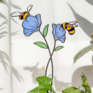 Peut inclure: Un pot en céramique blanc texturé avec des plantes vertes et deux piquets de jardin en forme de fleurs bleues avec des accents d'abeilles. Les piquets ont des tiges noires, des feuilles vertes et des pétales de fleurs bleues. Les abeilles sont jaunes et noires.