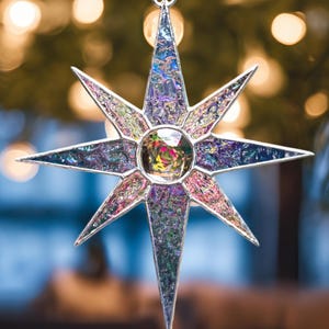 May include: A star-shaped Christmas ornament with a silver-colored metal frame and a colorful, iridescent glass design. The ornament has a central, round, clear gem and is suspended by a silver ribbon. The background is blurred with warm, out-of-focus lights.