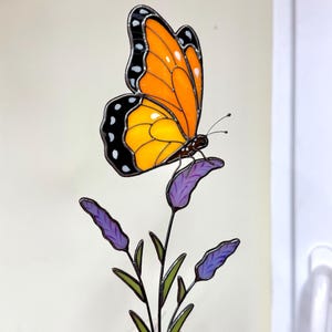 May include: A stained glass butterfly with orange and yellow wings, black edges, and white spots, perched on a purple flower. The butterfly is detailed with black antennae and sits atop a green stem with purple buds.