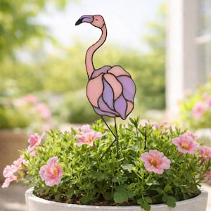 May include: A stained glass flamingo garden stake in pink and purple hues. The flamingo has a long, curved neck and stands on thin metal legs. It is placed in a pot of pink flowers, creating a decorative garden accent.