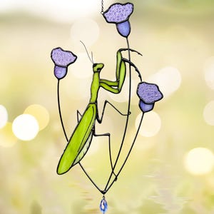 以下が含まれることがあります： 緑色のカマキリが紫色の花が咲いた茎にとまっているステンドグラスの吊り下げ飾り。カマキリは羽根と脚が細かく描かれている。花は様式化されており、質感がある。茎の下には青いクリスタルがぶら下がっている。