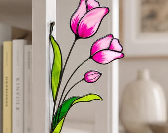 May include: A stained glass tulip decoration featuring three pink flowers with black stems and green leaves. The flowers are a vibrant pink with darker pink edges. The leaves are a bright green. The decoration is mounted on a white surface.
