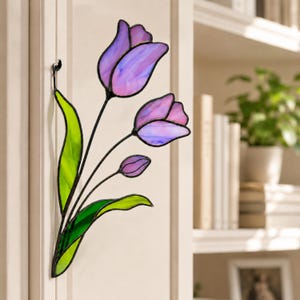 Peut inclure: Décoration de tulipes en vitrail avec deux fleurs épanouies et un bouton en nuances de violet et de rose. Les feuilles sont vertes et les tiges noires. L'œuvre est montée sur une surface blanche.
