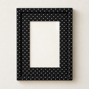 May include: A rectangular picture frame featuring a black fabric covering with a repeating white polka dot pattern. The frame has a thick, padded border surrounding a central white rectangular opening for displaying a photograph or artwork.