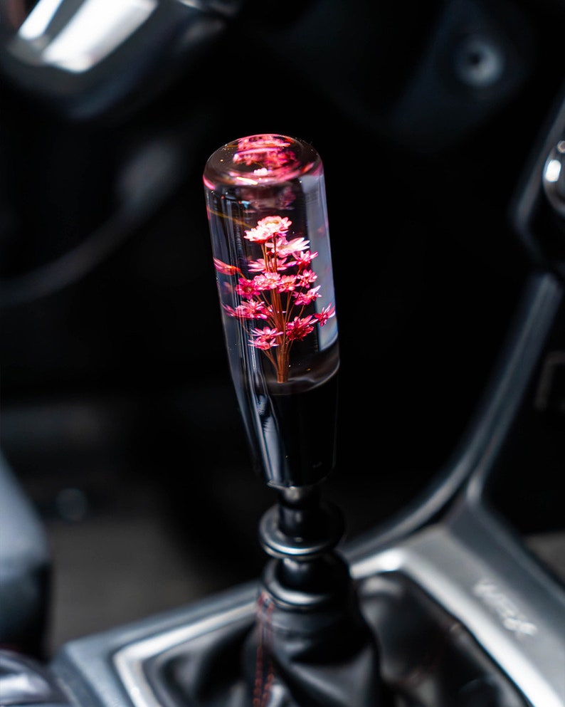 May include: A car gear stick knob with a clear, cylindrical top containing pink flowers. The base is black and the shift lever is visible in the background. The car's interior is dark.